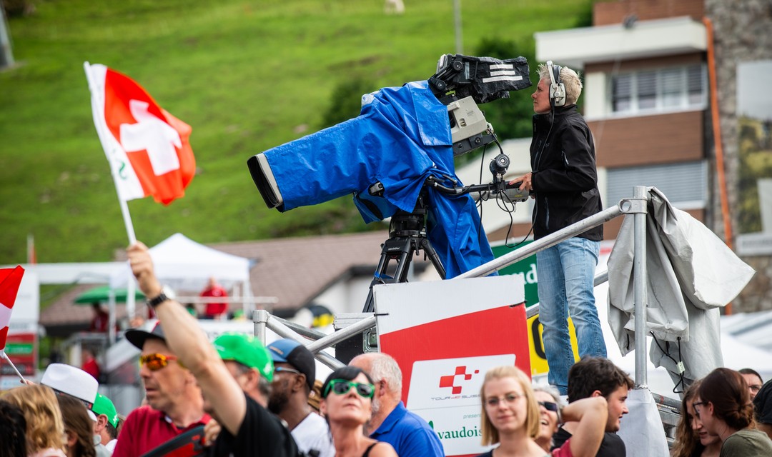 Kamera für Live-Übertragung auf Podest, dahinter Kamerafrau. Die Kamera ist eingepackt in blaue Folie gegen Regen. Im Vordergrund sind Fans, jemand schwenkt eine kleine Schweizer Fahne.