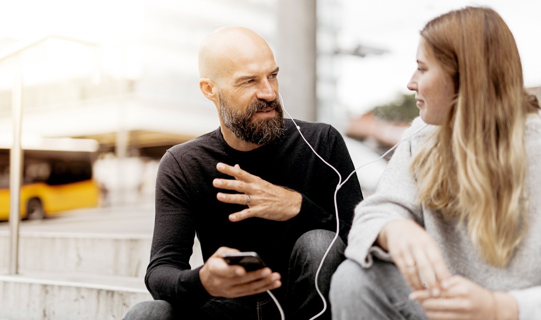 Mann mit Bart und Glatze hört Musik mit junger Frau, beide haben je einen Höhrer eines In-Ear-Kopfhörers im Ohr